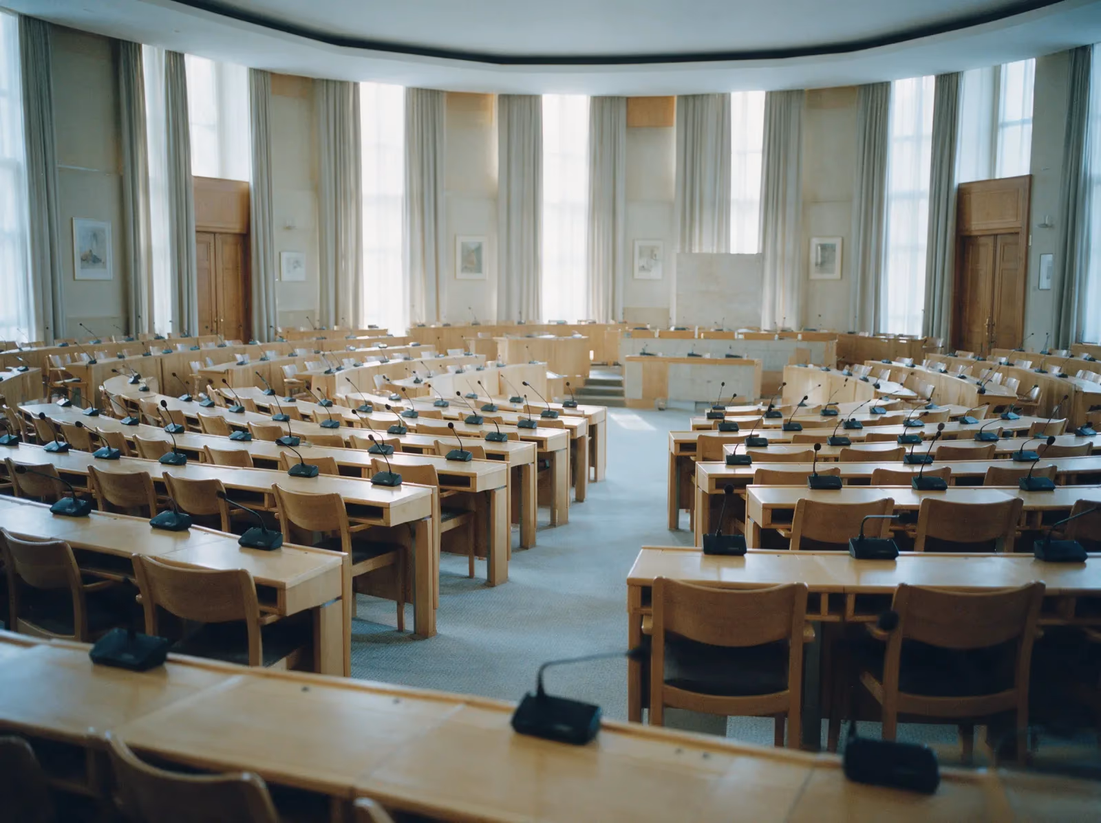 Curved rows of empty desks with microphones in a council assembly chamber, lit by tall windows