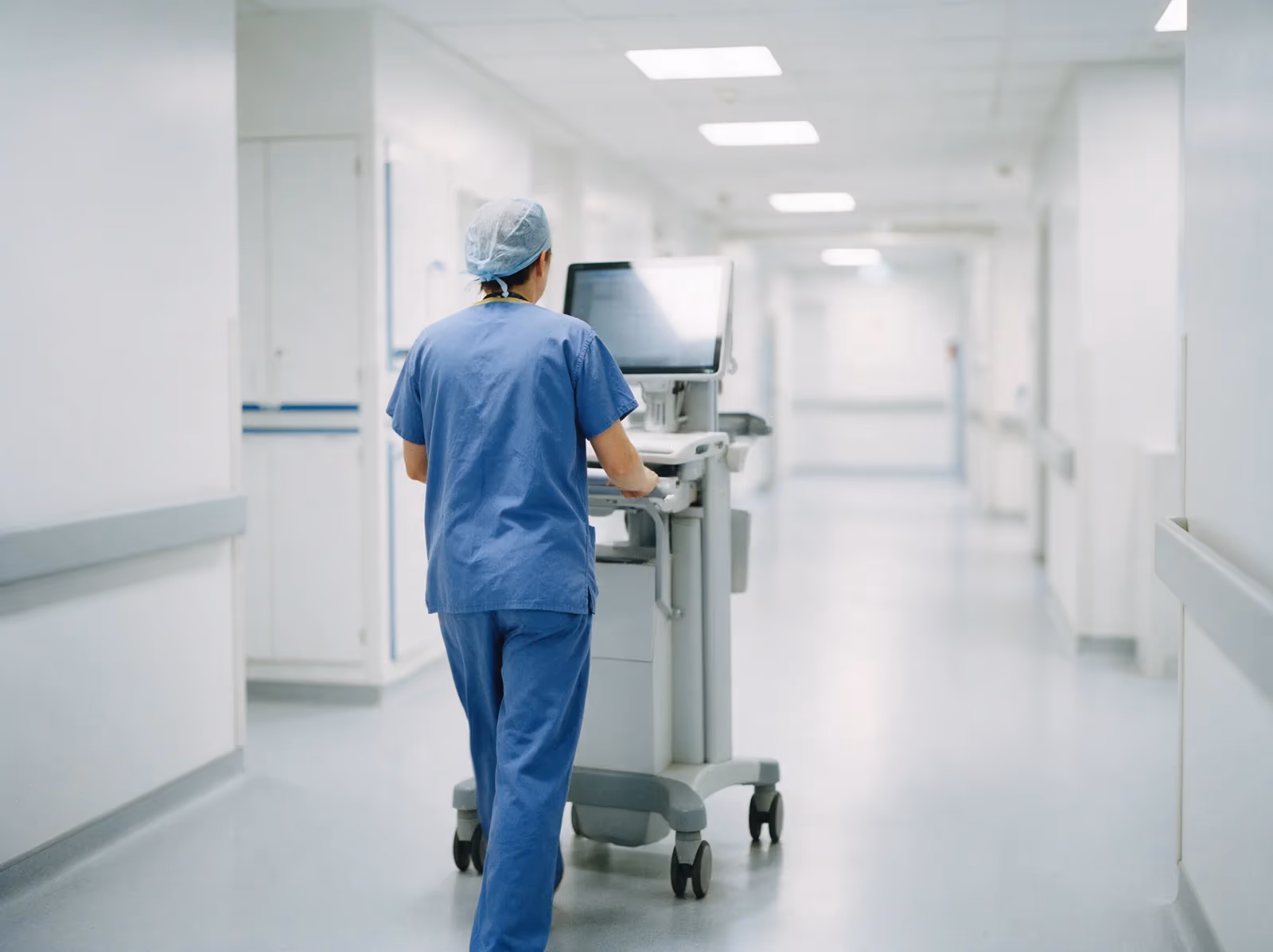 A clinician seen from behind pushing a mobile computer workstation along a bright modern hospital corridor