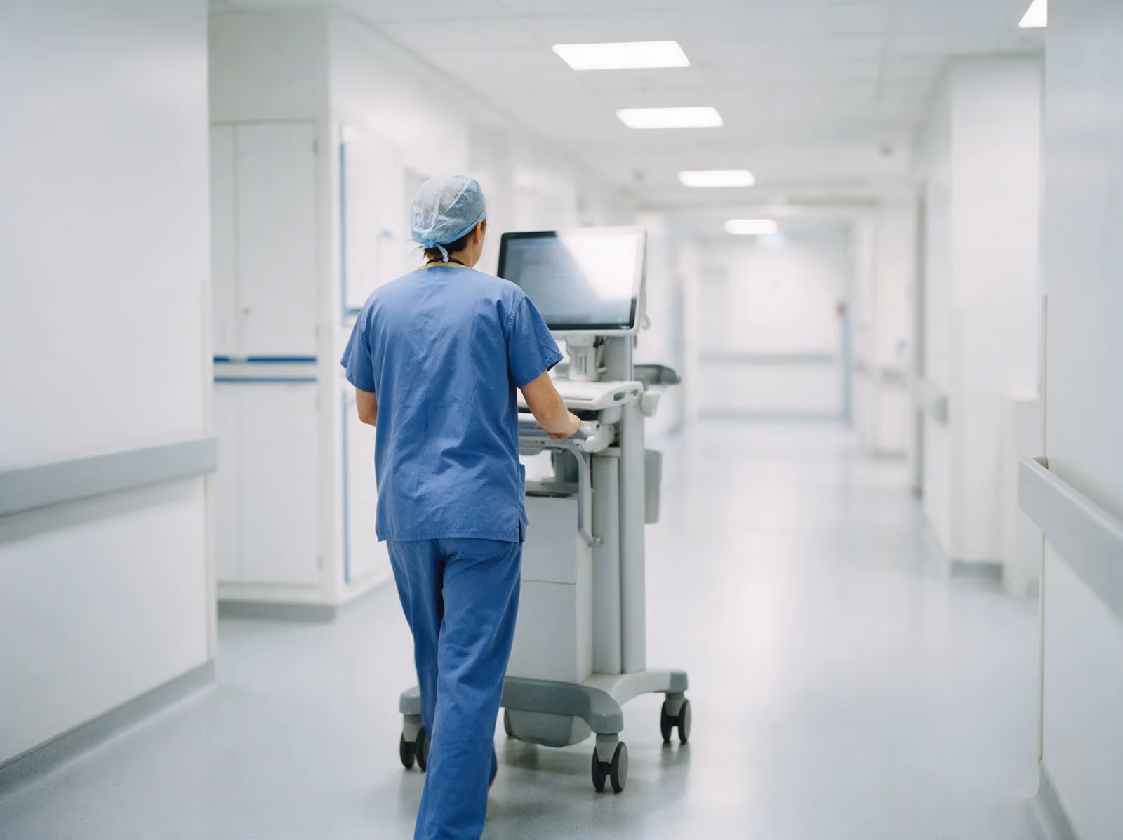 A clinician seen from behind pushing a mobile computer workstation along a bright modern hospital corridor